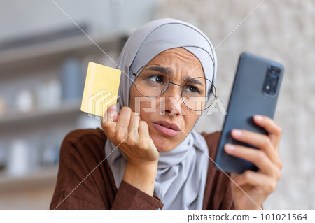 Close-up photo. The account is blocked, there are not enough funds in the account. Failed payment, transaction. A Muslim woman in hijab, holding a credit card in her hands, looking upset at the phone. 101021564