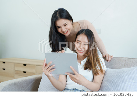 lgbtq, lgbt concept, homosexuality, portrait of two asian women posing happy together and loving each other while playing tablet at sofa. 101023483