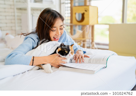 Asain woman lie on bed with read the book for multicolor cat that lie beside her in bedroom 101024762