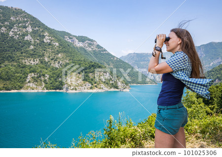 Young woman using binoculars exploring the nature at the Piva lake in Montenegro 101025306