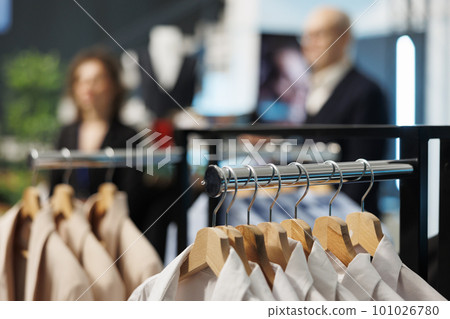 Selective focus of clothes on hangers in modern boutique, in background customers shopping for casual wear. Clients buying fashionable clothes and trendy accessories in clothing store Selective focus of clothes on hangers in modern boutique, in background customers shopping for casual wear. Clients buying fashionable clothes and trendy accessories in clothing store 101026780