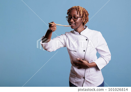 Female chef dressed in work uniform testing preparation of dish with wooden spoon in studio shot. African american expert cook delighting herself with a perfectly prepared recipe. 101026814