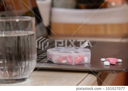 Organizer weekly shots with glass of water on workplace background and laptop Closeup of medical pill box with doses of tablets for daily take medicine with white pink drugs and capsules. Health care 101027077