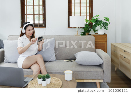 Portrait of asian woman using smart phone mobile and headphones relaxing on sofa at home. 101030121