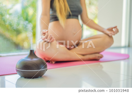 Young pregnant woman doing yoga exercises and meditating at home with an aroma diffuser. Health care, mindfulness, relaxation and wellness concept. Aromatherapy during pregnancy 101031948
