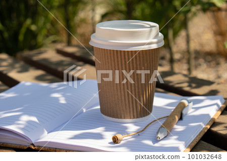 Eco recycling paper cup with coffee or tea on kraft paper with empty paper notebook on wooden bench. Concept of study work outdoors. Take away coffee to go. Copy space for text. Disposable Cardboard 101032648