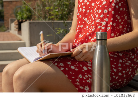Unrecognizable Young woman in red dress drinking water from metal bottle Writing Gratitude Journal on wooden bench. Today I am grateful for. Self discovery journal, self reflection creative writing 101032668
