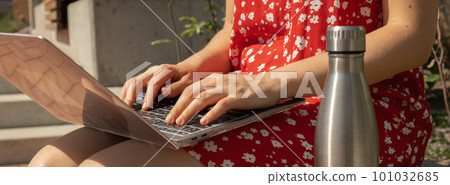 Unrecognizable Young woman in red dress drinking water from reusable metal bottle with laptop. Freelancer's place of work. Study and work online. Remote business education. View webinar. E-learning 101032685