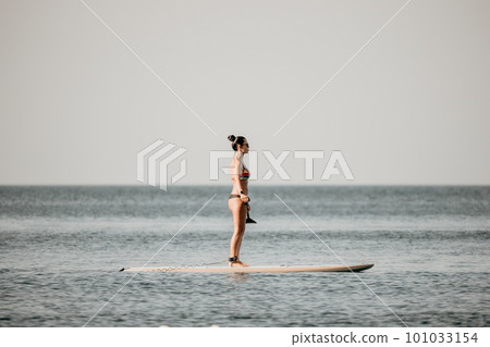 Sea woman sup. Silhouette of happy middle aged tanned woman in rainbow bikini, surfing on SUP board, confident paddling through water surface. Idyllic sunset. Active lifestyle at sea or river. Sea woman sup. Silhouette of happy middle aged tanned woman in rainbow bikini, surfing on SUP board, confident paddling through water surface. Idyllic sunset. Active lifestyle at sea or river. 101033154