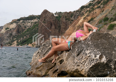 Woman travel sea. Pink bikini tourist captures sea memory, posing on beach amidst volcanic mountains for travel adventure. Happy woman enjoys outdoor photography. Woman travel sea. Pink bikini tourist captures sea memory, posing on beach amidst volcanic mountains for travel adventure. Happy woman enjoys outdoor photography. 101033208