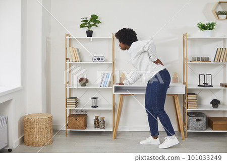 Young black woman feels severe back pain while at home in the living room. 101033249