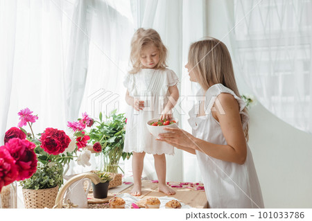 A little blonde girl with her mom on a kitchen countertop decorated with peonies. The concept of the relationship between mother and daughter. Spring atmosphere. A little blonde girl with her mom on a kitchen countertop decorated with peonies. The concept of the relationship between mother and daughter. Spring atmosphere. 101033786