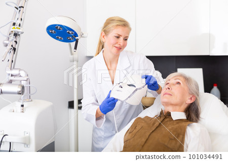 Cosmetologist using spectrum mask on elderly woman 101034491