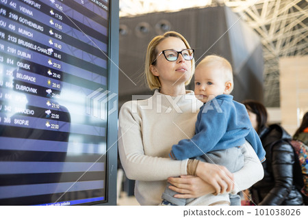 Mother traveling with child, holding his infant baby boy at airport terminal, checking flight schedule, waiting to board a plane. Travel with kids concept 101038026