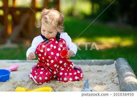 Cute toddler girl playing in sand on outdoor playground. Beautiful baby in red gum trousers having fun on sunny warm summer day. Child with colorful sand toys. Healthy active baby outdoors plays games Cute toddler girl playing in sand on outdoor playground. Beautiful baby in red gum trousers having fun on sunny warm summer day. Child with colorful sand toys. Healthy active baby outdoors plays games 101038450