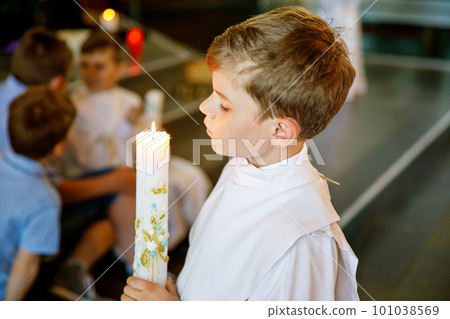 Little kid boy receiving his first holy communion. Happy child holding Christening candle. Tradition in catholic curch. Kid in a white traditional gown in a church near altar. Little kid boy receiving his first holy communion. Happy child holding Christening candle. Tradition in catholic curch. Kid in a white traditional gown in a church near altar. 101038569
