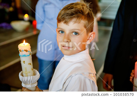 Little kid boy receiving his first holy communion. Happy child holding Christening candle. Tradition in catholic curch. Kid in a white traditional gown in a church near altar. 101038570