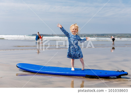Little cute toddler girl at the Ballybunion surfer beach, having fun on surfboard for the first time, west coast of Ireland. Happy child enjoying Irish summer with family. Little cute toddler girl at the Ballybunion surfer beach, having fun on surfboard for the first time, west coast of Ireland. Happy child enjoying Irish summer with family. 101038603