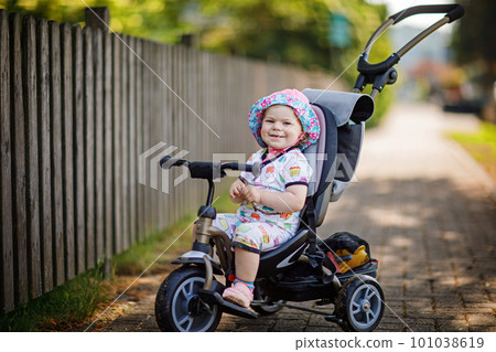 Cute adorable toddler girl sitting on pushing bicyle or tricycle. Little baby child going for a walk with parents on sunny day. Happy healthy kid in colorful clothes Cute adorable toddler girl sitting on pushing bicyle or tricycle. Little baby child going for a walk with parents on sunny day. Happy healthy kid in colorful clothes 101038619
