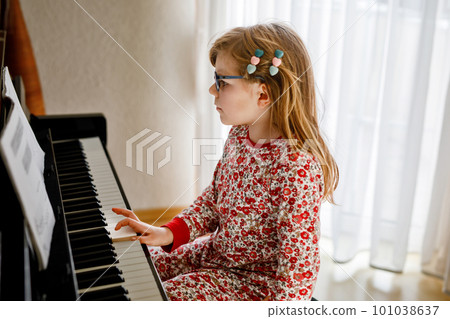 Little happy girl playing piano in living room. Cute preschool child with eye glasses having fun with learning to play music instrument. Little happy girl playing piano in living room. Cute preschool child with eye glasses having fun with learning to play music instrument. 101038637