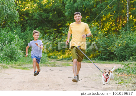 Father and son jogging along country dirt road with dog on waist leash 101039657