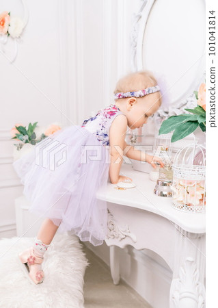 Baby girl elegant dress. A one-year-old girl in a puffy dress and a cute bow poses against the backdrop of a bright room with a dressing table and flowers. Baby girl elegant dress. A one-year-old girl in a puffy dress and a cute bow poses against the backdrop of a bright room with a dressing table and flowers. 101041814