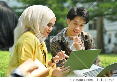 Smiling Muslim student and her friend using digital tablet on green meadow in campus. Education, youth and community concept 101042792