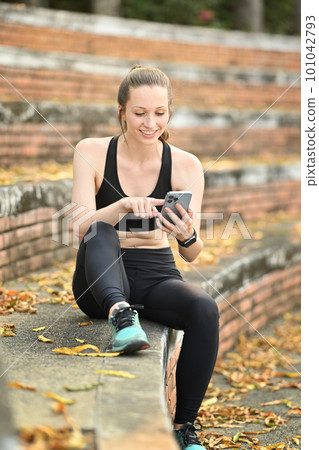 Beautiful caucasian female runner in sport clothes using smart phone, resting after morning workout Beautiful caucasian female runner in sport clothes using smart phone, resting after morning workout 101042793