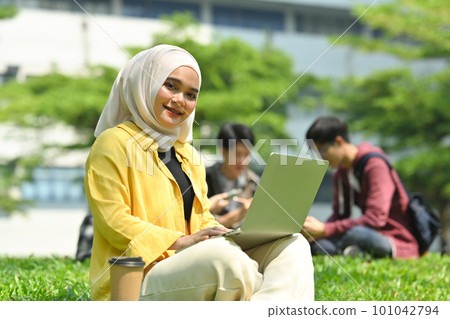 Smiling muslim college student using laptop on green grass in the campus. Education, technology and lifestyle concept 101042794