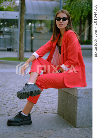 Blonde girl in a red lady-type pantsuit and black top, watch, pendant, sunglasses is posing sitting on a stone bench. Fashion concept. Full-length. Blonde girl in a red lady-type pantsuit and black top, watch, pendant, sunglasses is posing sitting on a stone bench. Fashion concept. Full-length. 101044316