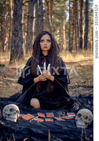 Witch in black dress with cape and hood. Posing in pine forest. Sitting on dark blanket whith fortune-telling cards and skulls on it. Full length. Witch in black dress with cape and hood. Posing in pine forest. Sitting on dark blanket whith fortune-telling cards and skulls on it. Full length. 101044324