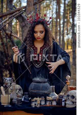 Witch in black, long dress, with red crown in her long hair. Posing in pine forest. Making a magic potion. Spells and witchcraft. Close-up, smoke. 101044329