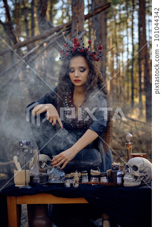 Witch in black, long dress, with red crown in her long hair. Posing in pine forest. Making a magic potion. Spells and witchcraft. Close-up, smoke. Witch in black, long dress, with red crown in her long hair. Posing in pine forest. Making a magic potion. Spells and witchcraft. Close-up, smoke. 101044342