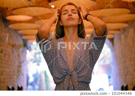 Blonde girl in long blue dress, watch, ring is posing standing under a huge chandeliers in a city cafe. Close-up shot. Blonde girl in long blue dress, watch, ring is posing standing under a huge chandeliers in a city cafe. Close-up shot. 101044346
