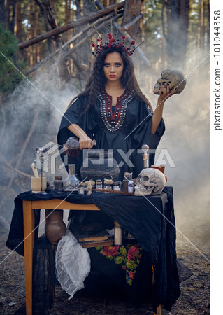 Witch in black, long dress, with red crown in her hair. Posing in pine forest, holding skull, making potion. Spells and witchcraft. Close-up, smoke. Witch in black, long dress, with red crown in her hair. Posing in pine forest, holding skull, making potion. Spells and witchcraft. Close-up, smoke. 101044358
