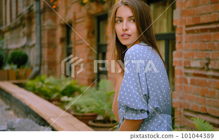 Blonde girl in long blue dress and a small black handbag on her shoulder is posing sitting sideways on a parapet. Fashion and style. Close-up shot. 101044389