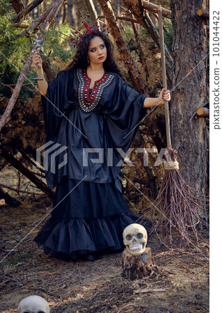 Witch in black, long dress, with red crown in her long hair. Posing with broom and skull in pine forest. Spells, magic and witchcraft. Full length. Witch in black, long dress, with red crown in her long hair. Posing with broom and skull in pine forest. Spells, magic and witchcraft. Full length. 101044422