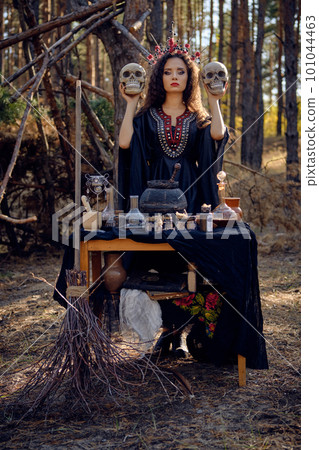 Witch in black, long dress, with red crown in her long hair. Posing in pine forest. Holding skulls. Spells, magic and witchcraft. Full length. Witch in black, long dress, with red crown in her long hair. Posing in pine forest. Holding skulls. Spells, magic and witchcraft. Full length. 101044463