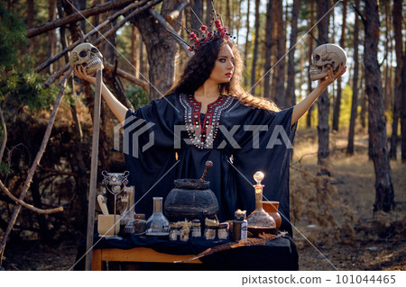 Witch in black, long dress, with red crown in her long, curly hair. Posing in pine forest. Holding skulls. Spells, magic and witchcraft. Close-up. Witch in black, long dress, with red crown in her long, curly hair. Posing in pine forest. Holding skulls. Spells, magic and witchcraft. Close-up. 101044465