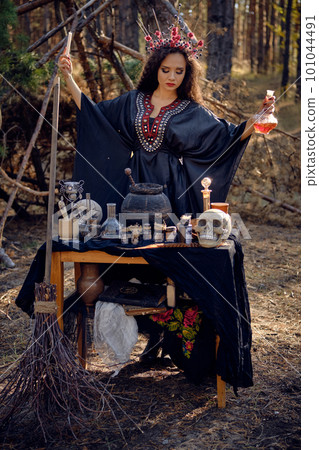 Beautiful witch in black, long dress, with red crown in her long hair. Posing in pine forest. Spells, magic and witchcraft. Full length portrait. 101044491