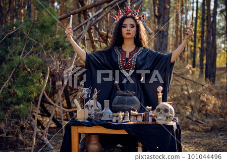 Beautiful witch in black, long dress, with red crown in her long, curly hair. Posing in pine forest. Spells, magic and witchcraft. Close-up. 101044496