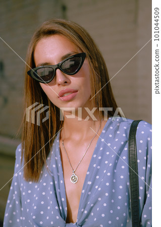 Blonde girl in long blue dress, sunglasses and a small black handbag on her shoulder is walking alone in the city. Fashion and style. Close-up shot. 101044509