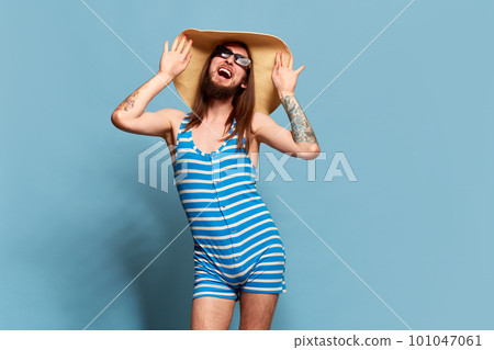 Portrait of young happy man in striped swimming suit, sunglasses and straw hat posing against blue studio background 101047061