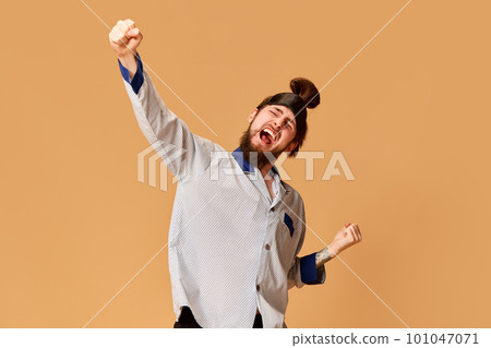 Portrait of young emotional man in gray pajamas and sleeping mask posing with shouting expression against orange studio background. Motivational energy 101047071