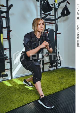 Beautiful blonde woman wearing in sensors and belts, fastens with velcro is practicing EMS fitness in a gym, doing squat. Modern kind of sport. Beautiful blonde woman wearing in sensors and belts, fastens with velcro is practicing EMS fitness in a gym, doing squat. Modern kind of sport. 101047497