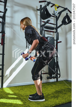 Beautiful blonde woman wearing in sensors and belts, fastens with velcro is practicing EMS fitness in a gym, building up muscles with a dumbbells. Beautiful blonde woman wearing in sensors and belts, fastens with velcro is practicing EMS fitness in a gym, building up muscles with a dumbbells. 101047507