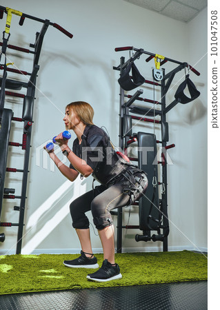 Beautiful blonde woman wearing in sensors and belts, fastens with velcro is practicing EMS fitness in a gym, building up muscles with a dumbbells. Beautiful blonde woman wearing in sensors and belts, fastens with velcro is practicing EMS fitness in a gym, building up muscles with a dumbbells. 101047508