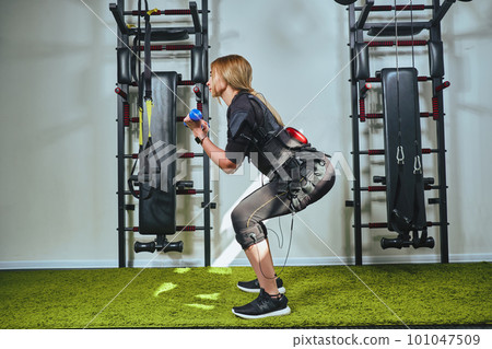 Beautiful blonde woman wearing in sensors and belts, fastens with velcro is practicing EMS fitness in a gym, building up muscles with a dumbbells. Beautiful blonde woman wearing in sensors and belts, fastens with velcro is practicing EMS fitness in a gym, building up muscles with a dumbbells. 101047509