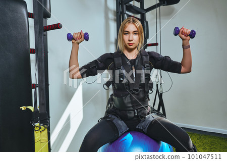 Girl wearing in sensors and belts, fastens with velcro practicing EMS fitness in gym, building up muscles with dumbbells, sitting on exercise ball. Girl wearing in sensors and belts, fastens with velcro practicing EMS fitness in gym, building up muscles with dumbbells, sitting on exercise ball. 101047511