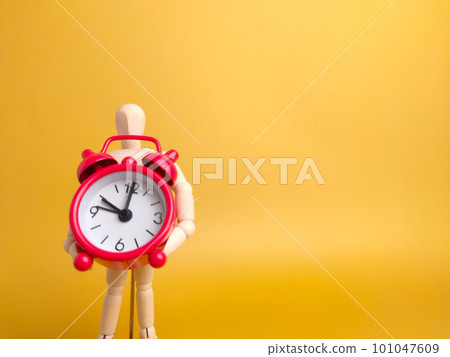 Wooden mannequin holding red alarm clock on a yellow background with copy space. 101047609